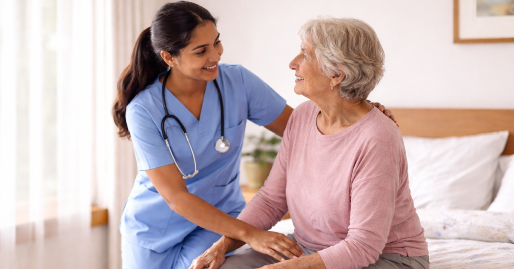 Young Nepali female nurse assisting a senior British woman to sit on the edge of the bed, demonstrating safe bed mobility care for elderly patients at home.