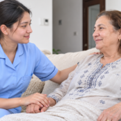 Young female Nepali nurse in a light blue uniform providing compassionate home nursing care to a 69-year-old Moroccan woman in a comfortable UAE home.