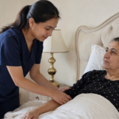 A professional home nurse in dark blue scrubs gently assisting an elderly female patient resting in bed in a home care setting.