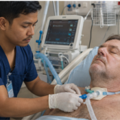 Young Nepali male nurse providing ventilator care to a 56-year-old German man at home, with the patient connected to a mechanical ventilator and resting comfortably in bed.