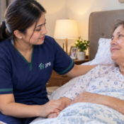 A home nurse in JPR Home Health Care uniform comforts a recovering elderly woman in bed while she receives oxygen support in a warm bedroom setting.