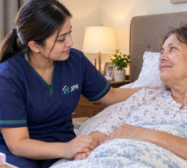 A home nurse in JPR Home Health Care uniform comforts a recovering elderly woman in bed while she receives oxygen support in a warm bedroom setting.